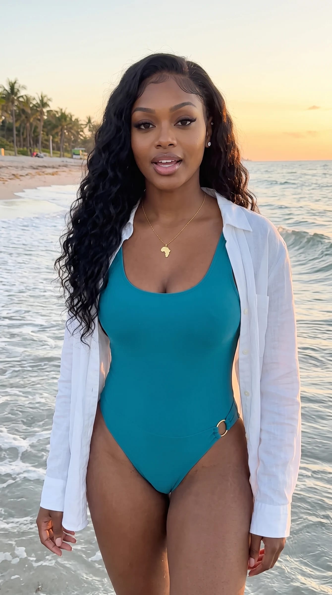 Imani at the waterline at golden hour in an ocean-teal one-piece and an open white linen shirt