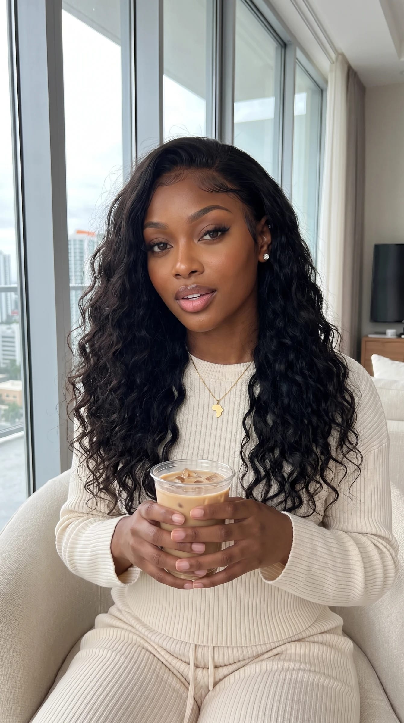 Imani in a cream knit lounge set by her window with an iced oat latte
